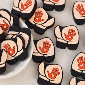May include: A collection of novelty butt-shaped pins. Each pin features a light skin-toned backside with black shorts and a red handprint. Some pins are in a white bowl.