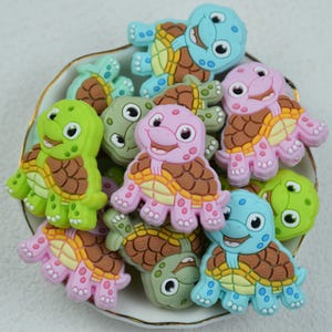 May include: A collection of colorful turtle-shaped embellishments in a white and gold-rimmed dish. The turtles are in shades of green, pink, blue, and brown. Each turtle has a smiling face and a detailed shell design.