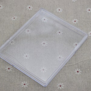 Puede incluir: Una caja rectangular de plástico transparente con un borde ligeramente elevado alrededor del perímetro. La caja está vacía y se encuentra sobre un fondo de tela marrón claro con un patrón floral blanco.