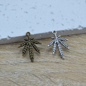 May include: Two metal leaf charms, one bronze and one silver, are shown against a neutral backdrop. The charms, with detailed veining and a loop for hanging, are suitable for jewellery or craft projects. The charms are approximately 3 cm in length.