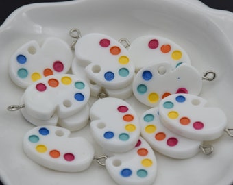 Puede incluir: Encantos de paleta de pintura blanca con puntos de pintura coloridos en rojo, naranja, amarillo, verde y azul. Cada dije tiene un lazo plateado para sujetar a joyas u otras manualidades. Los dijes están en un plato blanco.