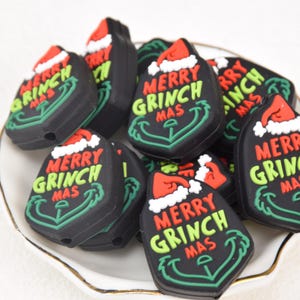 Könnte beinhalten: Eine Ansammlung schwarzer, tropfenförmiger Perlen mit dem Text "MERRY GRINCH MAS" in Grün und Rot. Jede Perle zeigt einen Weihnachtsmannhut und ein grünes Ankerdesign, ideal für Weihnachtsdekorationen.