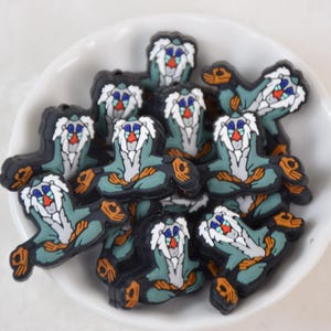 May include: A collection of cartoon-style rubber charms in a white bowl. Each charm features a character with a white beard, blue eyes, and a red nose, with a teal body and black outline.