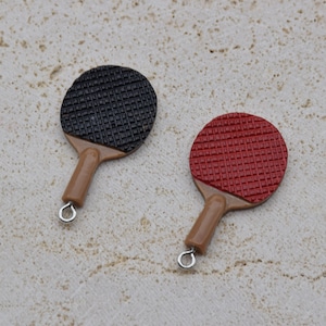 May include: Two miniature table tennis paddles, one with a black surface and one with a red surface. Both paddles have brown wooden handles.