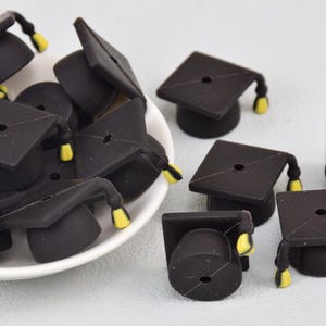 Op de afbeelding: Een groep zwarte, siliconen graduation cap-charms met gele kwastjes. De charms liggen verspreid op een wit oppervlak.