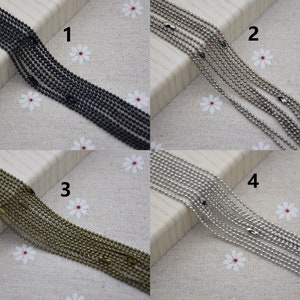 May include: Four strands of ball chain in different finishes: black, silver, bronze, and nickel. The chains are laid out on a white fabric background with small white flowers.