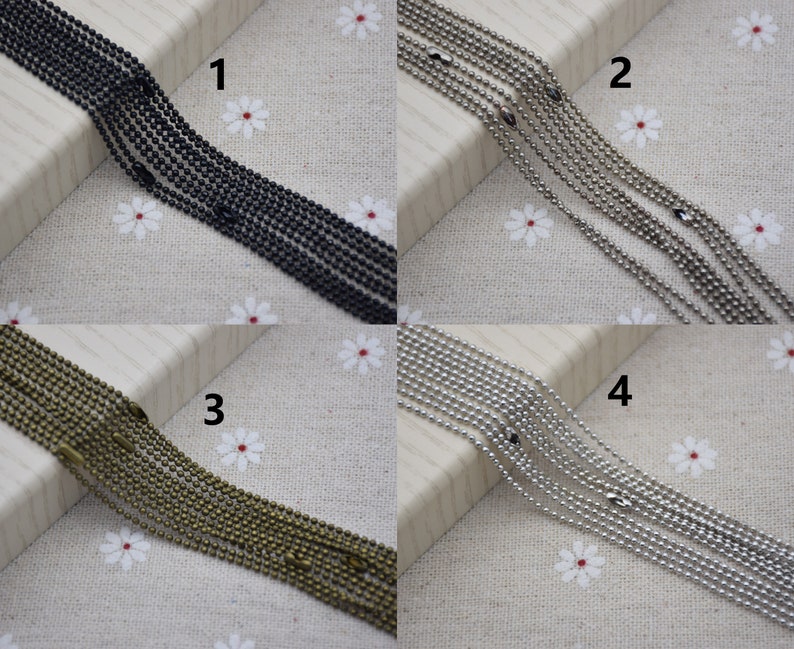 May include: Four strands of ball chain in different finishes: black, silver, bronze, and nickel. The chains are laid out on a white fabric background with small white flowers.