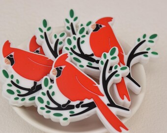 Può includere: Una collezione di ornamenti a forma di uccello cardinale rosso con becchi e accenti neri, appollaiati su rami bianchi con foglie verdi. Gli uccelli sono disposti in un piatto bianco.