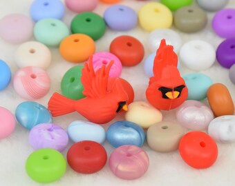 Può includere: Una collezione di perline in silicone colorate con due figurine di uccelli cardinali rossi. Le perline sono di varie forme e dimensioni, tra cui rotonde, ovali e a forma di ciambella.
