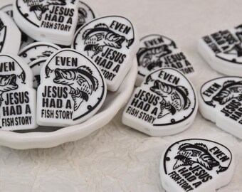 May include: White, heart-shaped beads with black text and a fish graphic. The text reads "EVEN JESUS HAD A FISH STORY". The beads are displayed in a white dish and scattered on a textured surface.