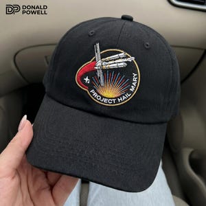 Puede incluir: Una gorra de béisbol negra con un parche circular bordado en la parte delantera. El parche muestra una nave espacial blanca con una estela roja, un resplandor solar amarillo y el texto PROJECT HAIL MARY en letras mayúsculas blancas.