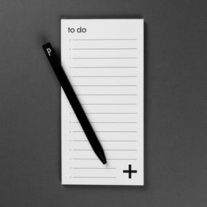 Op de afbeelding: Een zwarte pen ligt op een wit notitieblok met de woorden "to do" bovenaan. Het notitieblok heeft horizontale lijnen en een plusteken onderaan. De pen heeft de letter "P" erop. De achtergrond is effen grijs.