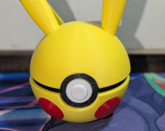 May include: A yellow and black Pikachu-themed Poké Ball. The ball has red accents and black detailing, with Pikachu's ears extending upwards. The design is a 3D-printed item, likely a collectible or toy.