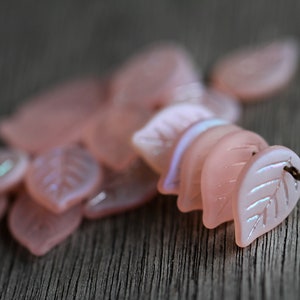 May include: A pile of pink iridescent glass leaf-shaped beads. The beads have a delicate, translucent quality and are perfect for jewelry making or crafting.