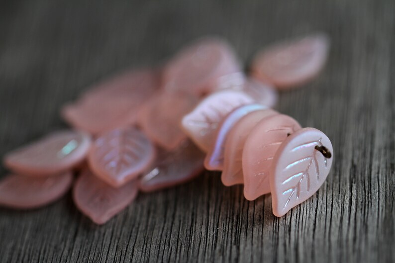 May include: A pile of pink iridescent glass leaf-shaped beads. The beads have a delicate, translucent quality and are perfect for jewelry making or crafting.