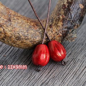 Könnte beinhalten: Zwei rote, tropfenförmige Perlen mit strukturierter Oberfläche und einer Metallschlaufe. Die Perlen sind etwa 1,1 cm x 0,9 cm groß. Sie werden vor einem Holzgrund gezeigt.