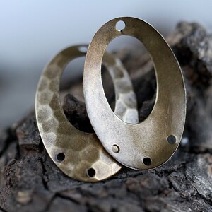 K&ouml;nnte beinhalten: Zwei ovale Metall-Anh&auml;nger mit geh&auml;mmerter Textur und einem Loch oben. Die Anh&auml;nger sind messingfarben und haben eine patinierte Oberfl&auml;che.
