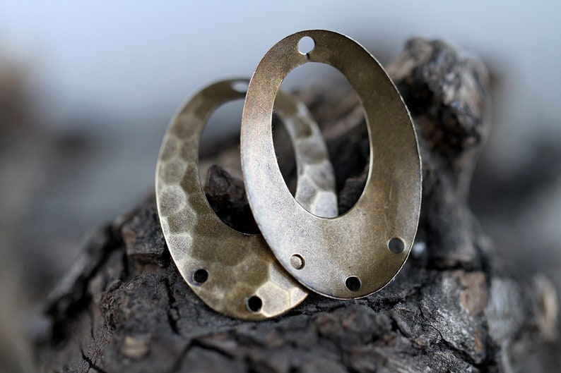 K&ouml;nnte beinhalten: Zwei ovale Metall-Anh&auml;nger mit geh&auml;mmerter Textur und einem Loch oben. Die Anh&auml;nger sind messingfarben und haben eine patinierte Oberfl&auml;che.