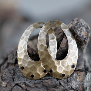 K&ouml;nnte beinhalten: Zwei ovale, metallene Ohrringrohlinge mit geh&auml;mmerter Textur und drei L&ouml;chern auf jeder Seite. Die Rohlinge sind messingfarben und liegen auf einem St&uuml;ck Holz.