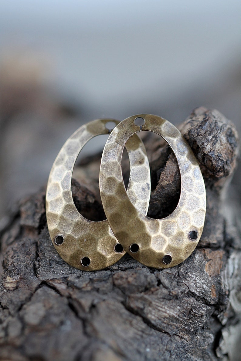 K&ouml;nnte beinhalten: Zwei ovale, metallene Ohrringrohlinge mit geh&auml;mmerter Textur und drei L&ouml;chern auf jeder Seite. Die Rohlinge sind messingfarben und liegen auf einem St&uuml;ck Holz.