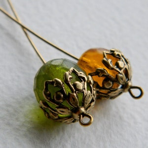May include: Two gold-toned metal bead caps with ornate floral designs. One bead cap is filled with a green glass bead, and the other is filled with an amber glass bead.