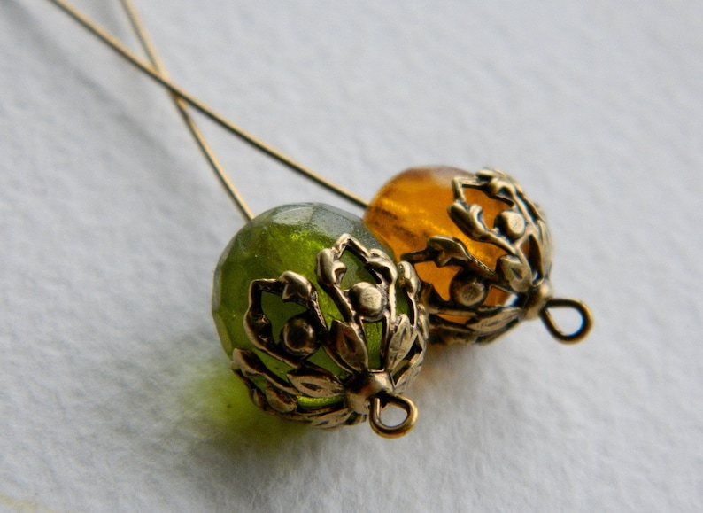 May include: Two gold-toned metal bead caps with ornate floral designs. One bead cap is filled with a green glass bead, and the other is filled with an amber glass bead.