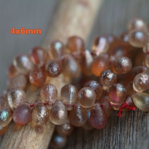 Könnte beinhalten: Eine Strähne aus irisierenden braunen Glasperlen, jede Perle ist etwa 4x6mm groß. Die Perlen sind auf einem dünnen roten Faden aufgefädelt und liegen auf einer Holzoberfläche.