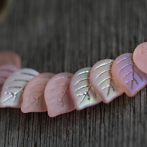 May include: A row of pink glass leaf-shaped beads with iridescent accents. The beads are arranged in a curved line on a wooden surface.