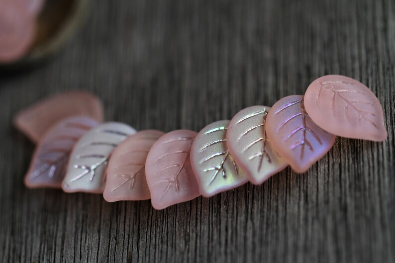 May include: A row of pink glass leaf-shaped beads with iridescent accents. The beads are arranged in a curved line on a wooden surface.