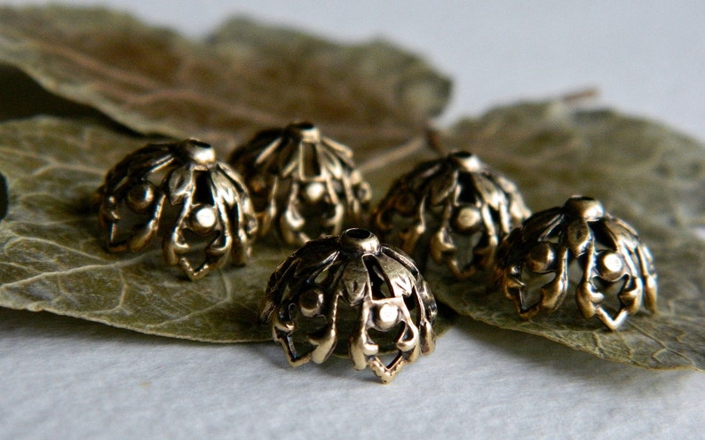 May include: Five gold-toned metal bead caps with a decorative, intricate design. The caps are arranged on a brown leaf.