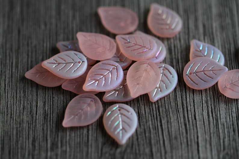 May include: A collection of pink glass leaf-shaped beads with a iridescent finish. The beads have a delicate etched design and are perfect for jewelry making or crafting.