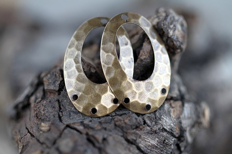K&ouml;nnte beinhalten: Zwei ovale Metall-Anh&auml;nger mit geh&auml;mmerter Textur und einem Loch in der Mitte. Die Anh&auml;nger sind messingfarben und haben eine patinierte Oberfl&auml;che.