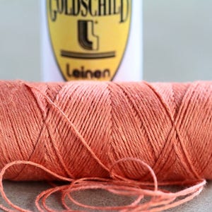 May include: A spool of orange linen thread with a label that reads "Goldschild Leinen".