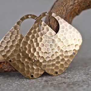 Puede incluir: Dos piezas en blanco para pendientes de metal dorado con textura martillada y forma cuadrada redondeada. Cada pieza en blanco tiene un agujero en el centro y tres agujeros alrededor del borde.