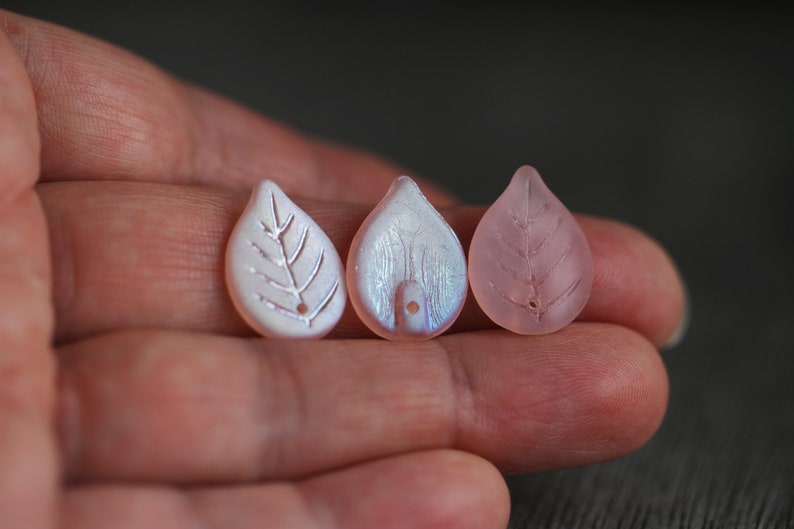 May include: Three leaf-shaped beads, one white with a silver leaf design, one iridescent white, and one pink.