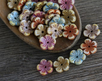Puede incluir: Surtido de cuentas de vidrio con forma de flor de colores con acentos metálicos. Las cuentas están dispuestas en una cuchara de madera.