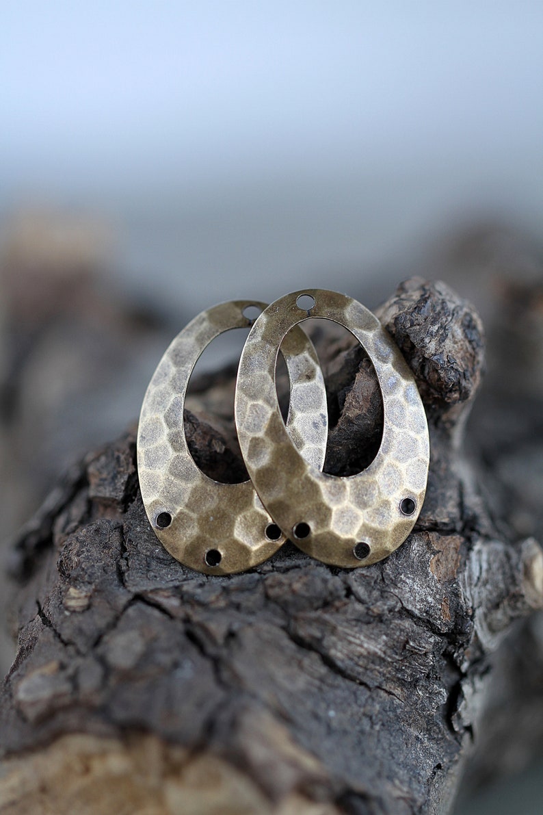 K&ouml;nnte beinhalten: Zwei ovale Metall-Anh&auml;nger mit geh&auml;mmerter Textur und Messing-Finish. Jeder Anh&auml;nger hat vier kleine L&ouml;cher am Rand.