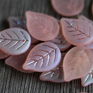 May include: A group of pink glass leaf-shaped beads with a frosted finish and a subtle iridescent sheen. The beads have a delicate leaf design etched into their surface.