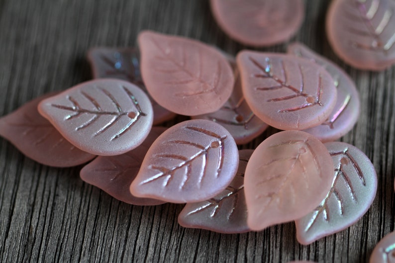 May include: A group of pink glass leaf-shaped beads with a frosted finish and a subtle iridescent sheen. The beads have a delicate leaf design etched into their surface.