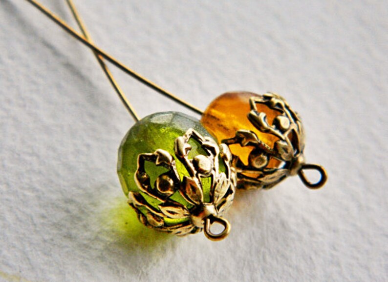 May include: Two gold-toned metal bead caps with intricate floral designs. One bead cap is green and the other is amber.