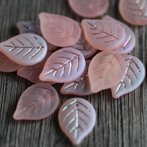 May include: A collection of pink glass leaf-shaped beads with a subtle iridescent shimmer. The beads have a delicate vein pattern etched into their surface.