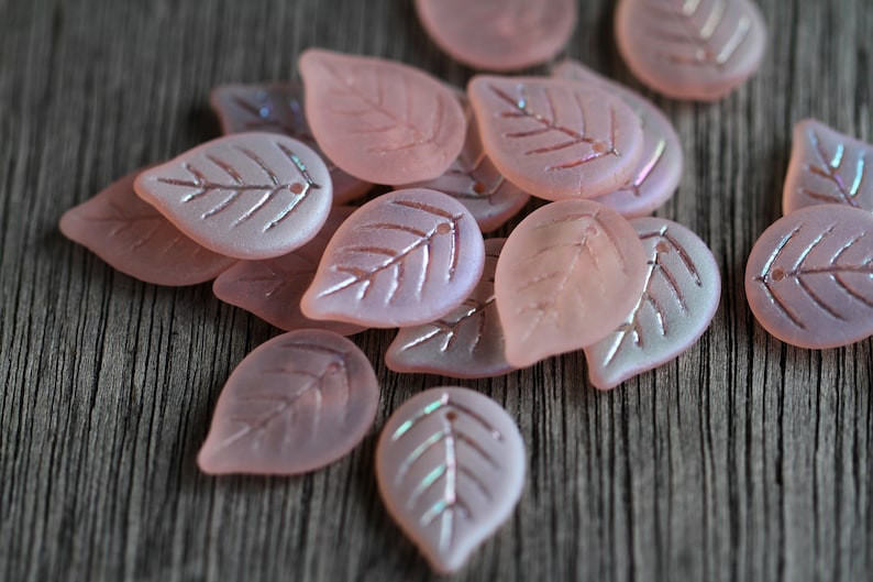 May include: A collection of pink glass leaf-shaped beads with a subtle iridescent shimmer. The beads have a delicate vein pattern etched into their surface.