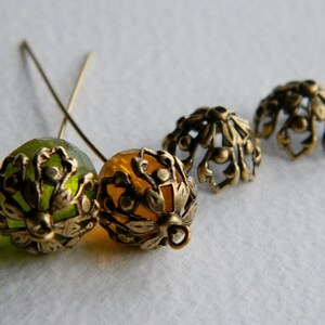 May include: Four decorative metal bead caps, two green and two gold, with intricate floral designs. The caps are attached to small metal pins.