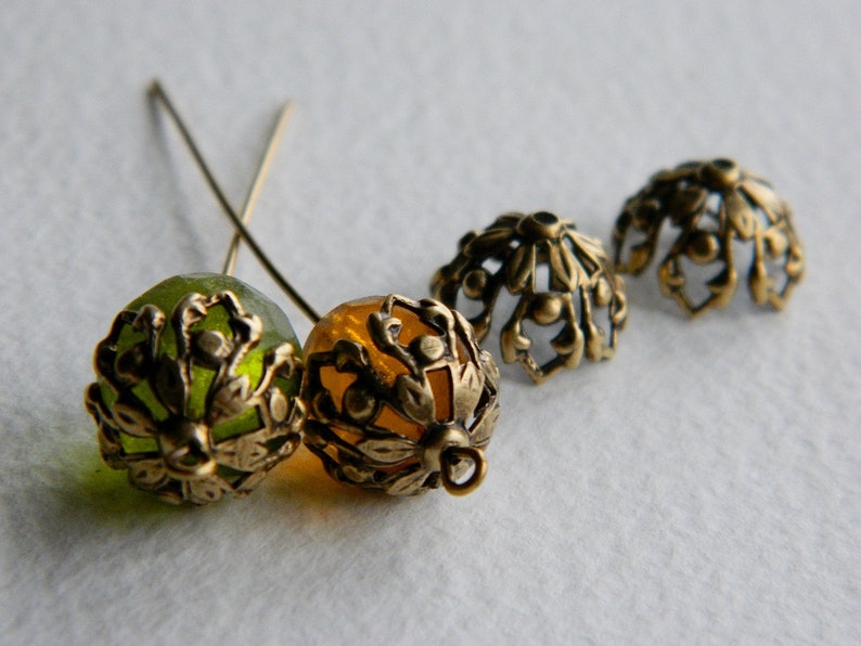 May include: Four decorative metal bead caps, two green and two gold, with intricate floral designs. The caps are attached to small metal pins.