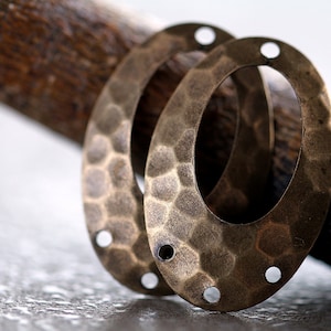 K&ouml;nnte beinhalten: Zwei ovale Metall-Anh&auml;nger mit geh&auml;mmerter Textur und Messing-Finish. Jeder Anh&auml;nger hat vier L&ouml;cher am Rand.