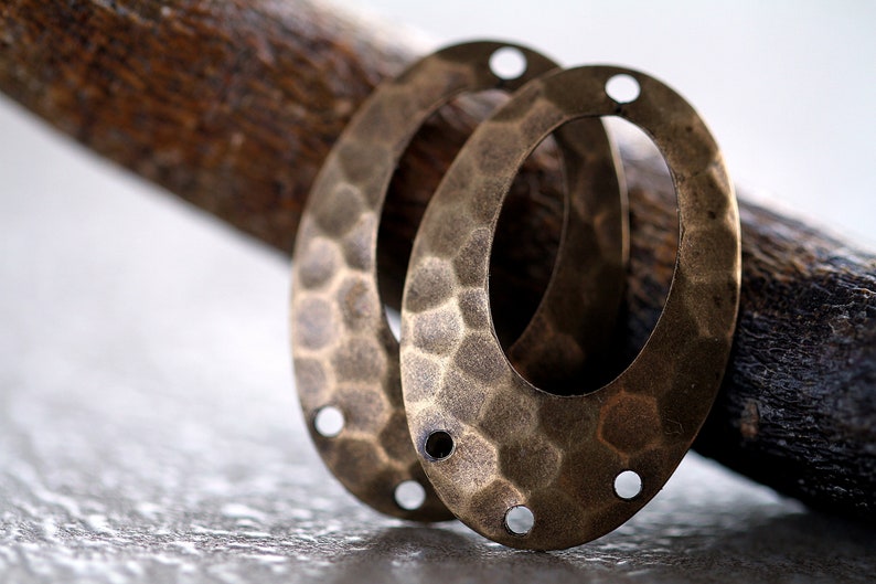 K&ouml;nnte beinhalten: Zwei ovale Metall-Anh&auml;nger mit geh&auml;mmerter Textur und Messing-Finish. Jeder Anh&auml;nger hat vier L&ouml;cher am Rand.