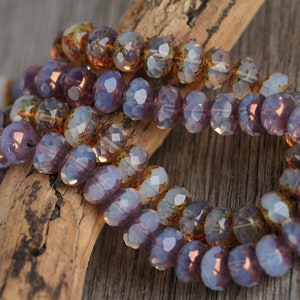 May include: Strands of faceted beads in shades of purple, amber, and clear. The beads are strung together and arranged on a piece of driftwood. The beads have a reflective, iridescent quality, and are likely used for jewelry making.