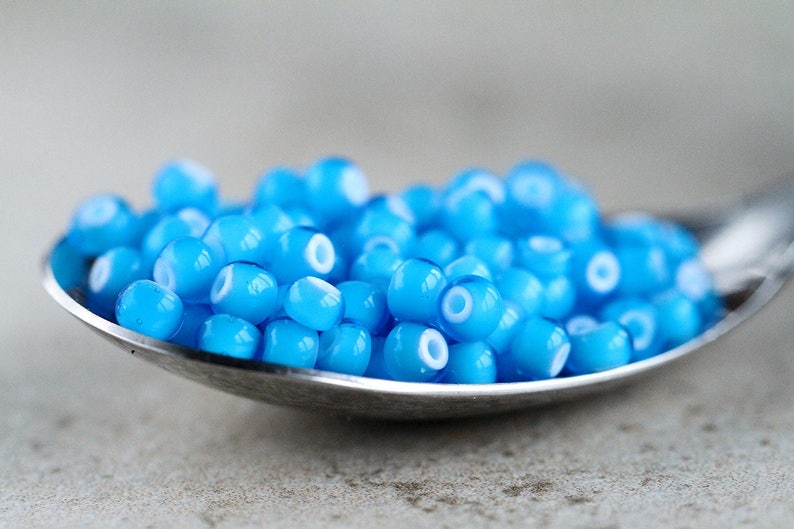 Puede incluir: Una cuchara llena de peque&ntilde;as cuentas de vidrio azul claro con un anillo blanco alrededor del centro de cada cuenta.