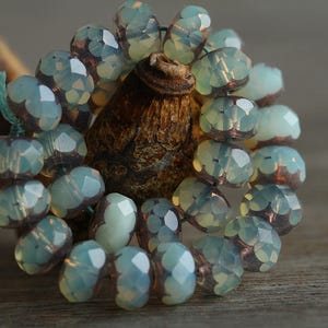 May include: Close-up of a strand of faceted, pale blue-green beads with a metallic sheen. The beads are strung together, forming a circular shape. The beads are set against a blurred, natural wood background.