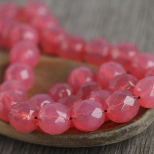 May include: A strand of pink faceted gemstone beads. The beads are strung on a clear elastic cord and are displayed on a wooden spoon.